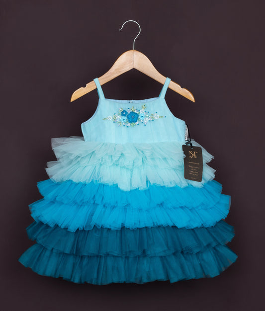 A sky blue tulle net frock with blue flowers and small pearls, displayed on a wooden hanger.