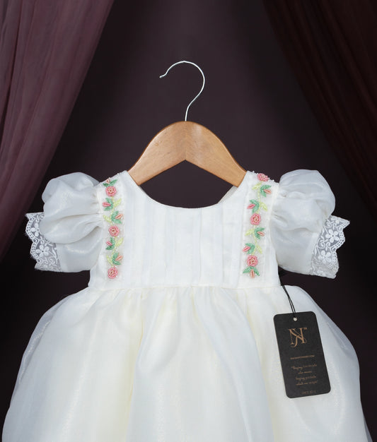 Close view of an off-white baby girl dress with floral embroidery, puff sleeves, and lace hem on a hanger.