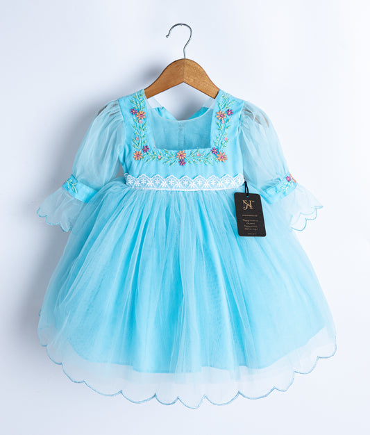 A blue below knee frock with floral embroidery on the yoke and lace-trimmed waistline, with a flowy skirt and scalloped end, displayed on a wooden hanger.