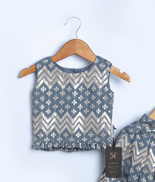 Close view of a stylish sleeveless kids blouse in a blue-grey shade, featuring gold zigzag and floral patterns with a subtle sheen. The blouse has a round neckline and a ruffled hem, displayed on a wooden hanger against a light background. A black brand tag hangs from the matching garment beside it.