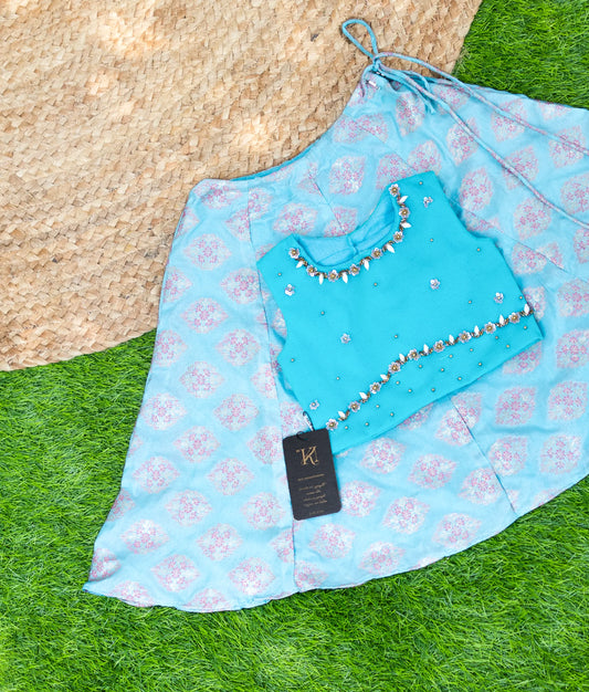 A vibrant traditional outfit laid out on grass and a woven mat. The set includes a turquoise blouse adorned with mirror work and a matching blue and pink patterned skirt with a drawstring waist. A black brand tag is attached to the skirt.