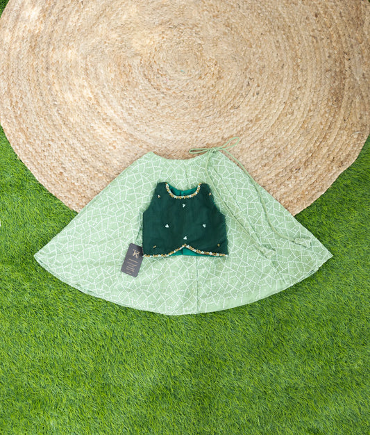 A festive green outfit featuring a sleeveless dark green embroidered top paired with a light green patterned skirt, displayed on grass with a jute mat background.