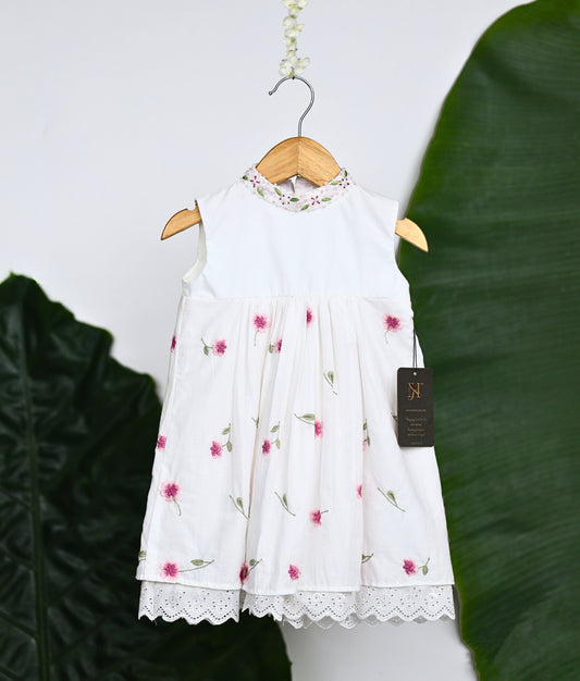 Sleeveless white baby dress with pink floral embroidery, lace hem, and embroidered neckline, hanging on a wooden hanger against a leafy green background.