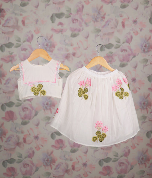White top and skirt set with pink floral embroidery and lace trim on wooden hangers.