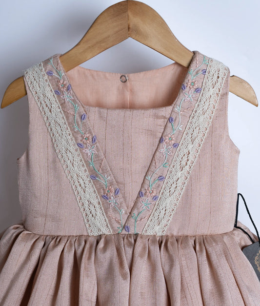 Close view of an beige, sleeveless baby frock with embroidered straps and a flared silhouette, displayed on a hanger.