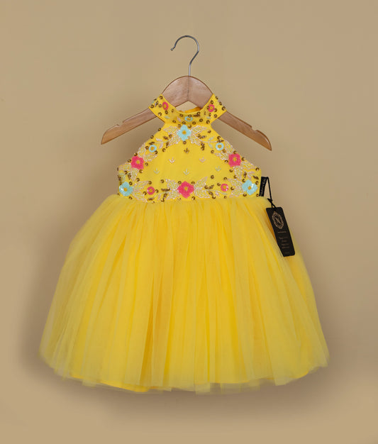 A yellow tulle net baby girl frock with floral handwork on the bodice, a halter neck, and a high neck closed at the back with a press button and tie, displayed on a hanger.