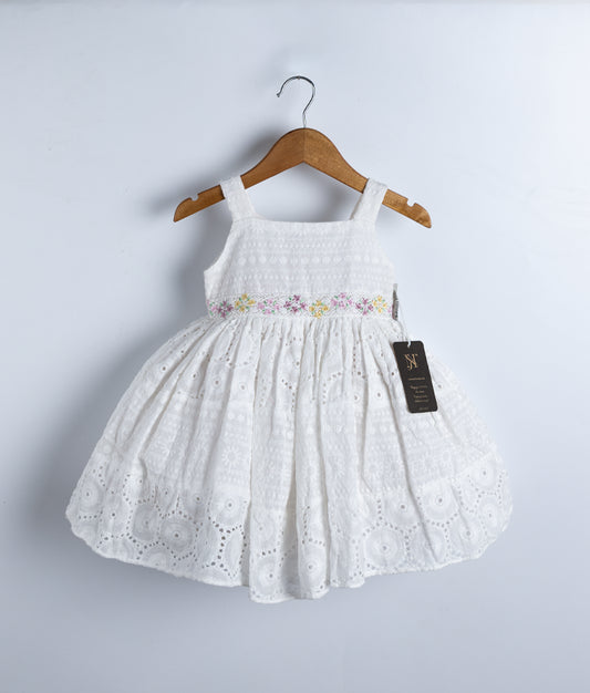 White sleeveless frock with lace belt and embroidery, hanging on a wooden hanger.