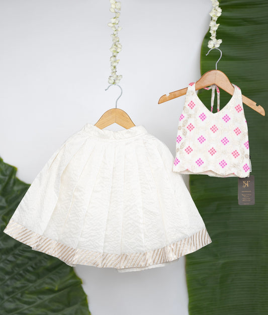 A traditional ethnic outfit featuring a white pleated skirt with a gold border and a white halter blouse adorned with pink and gold motifs, displayed against banana leaves and floral hangers.