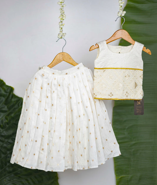 A traditional white and gold sleeveless blouse with embroidered detailing paired with a white pleated skirt featuring gold polka dots—a festive ethnic outfit for girls displayed on hangers against a leafy background.
