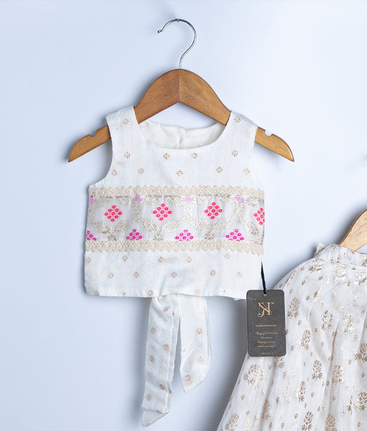 An ivory sleeveless baby girl crop top with gold and pink mirror embroidery, paired with a matching lehenga, displayed on a wooden hanger against a light background.