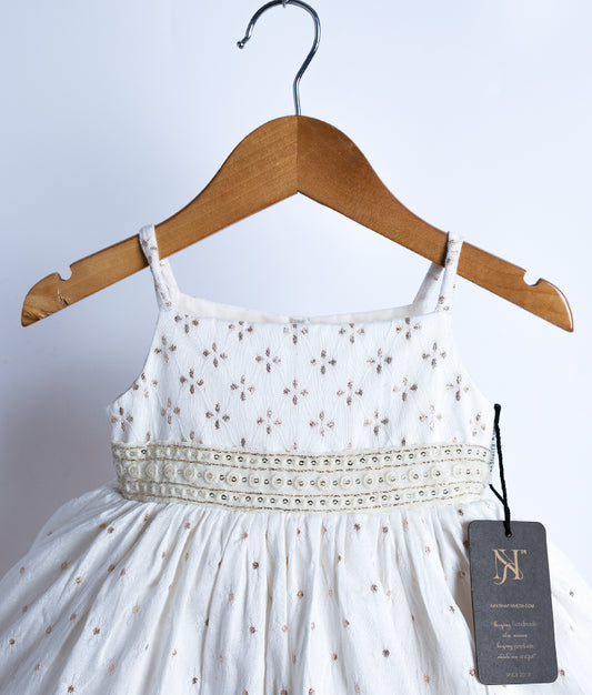 Close view of an off-white, sleeveless, flared frock displayed on a wooden hanger against a white background.