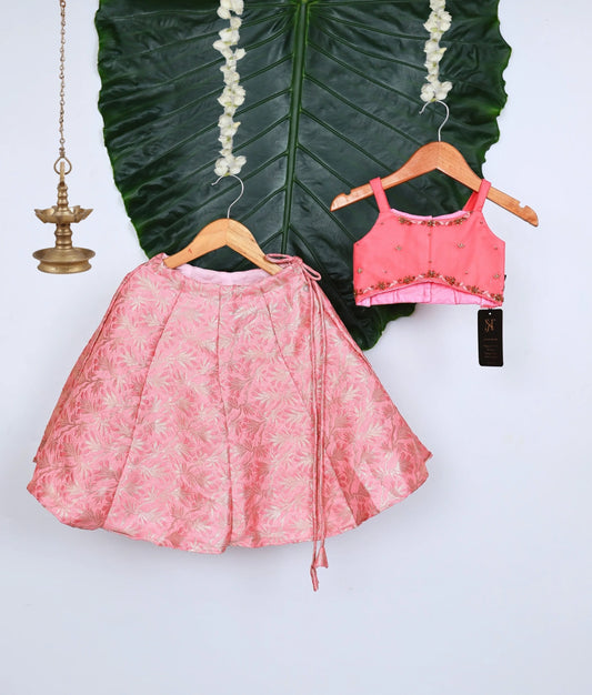 Stylish pink kids lehenga set with floral brocade skirt and embellished sleeveless top, displayed on hangers against a large green leaf backdrop with decorative brass lamp.