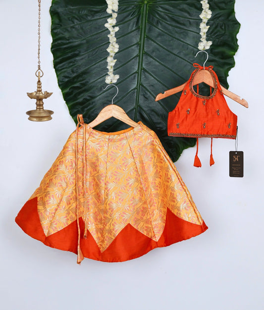 Stylish baby girl outfit with orange and gold brocade skirt and matching embellished halter neck blouse, displayed with traditional festive decor