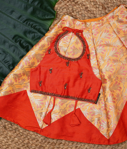Close-up of a stylish baby girl outfit with orange and gold brocade skirt and matching embellished halter neck blouse, displayed with traditional festive decor