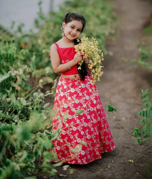 A girl wearing a red and pink floral tulle net top with a chanderi skirt standing in a outdoor setting with greenery in the background.