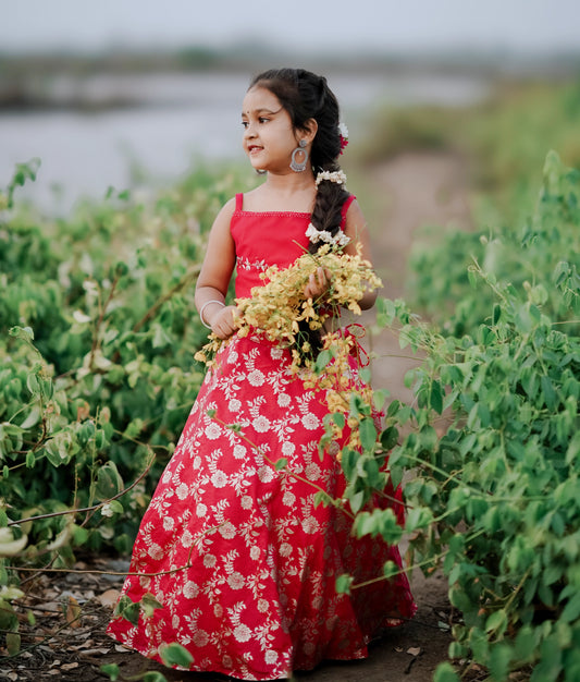 Young girl wearing a festive red chanderi skirt with golden floral embroidery, posing outdoors in a green field while holding yellow flowers.