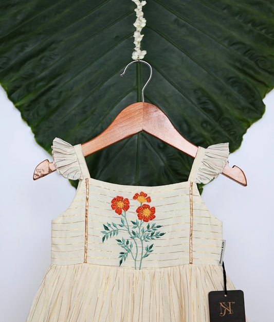 Close view of an off-white pleated frock with floral embroidery and ruffled sleeves and hem, displayed on a hanger.