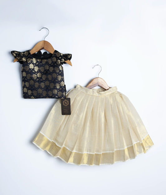 Elegant baby girl lehenga choli set featuring black brocade blouse with gold floral motifs and ruffle sleeves, paired with cream-gold silk pleated skirt, displayed on wooden hangers against white background
