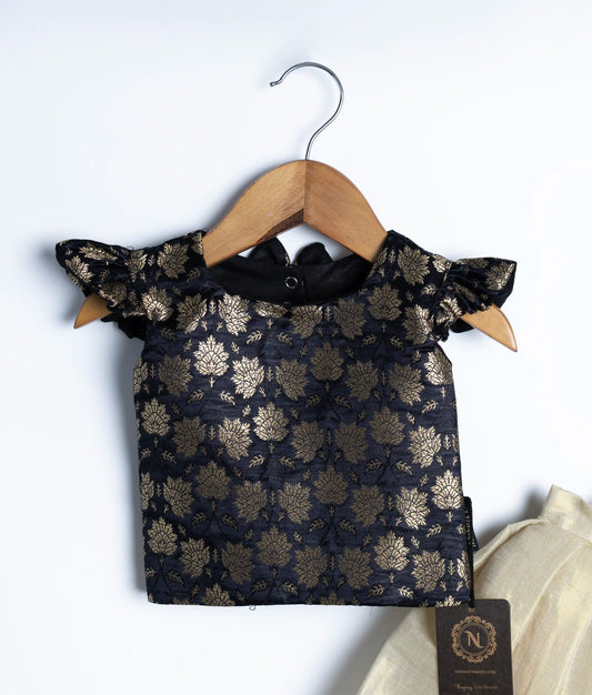 A close view of elegant baby girl lehenga choli set featuring black brocade blouse with gold floral motifs and ruffle sleeves, paired with cream-gold silk pleated skirt, displayed on wooden hangers against white background