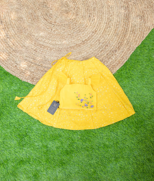 A yellow festive clothing set featuring a floral embroidered crop top with straps and a matching flared skirt with eyelet detailing, displayed on grass and a jute mat background.