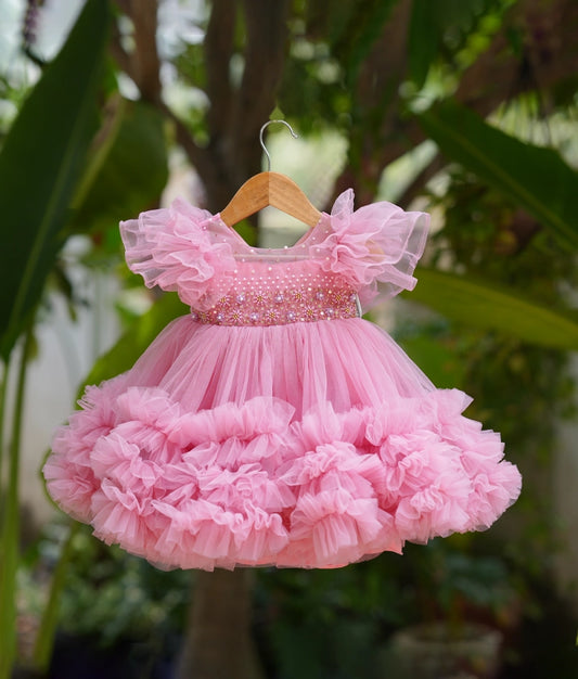 Pink ruffled birthday frock for baby girls with pearl-studded bodice, perfect for special occasions—displayed on hanger outdoors