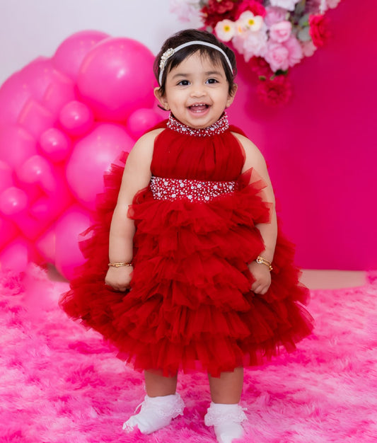 A baby girl in a maroon tulle net frock with handwork on the neck and waist, standing in front of a pink backdrop with balloons and flowers.
