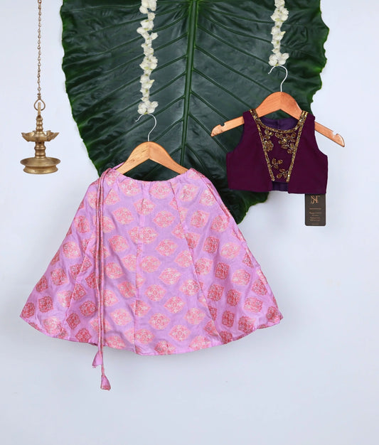 A traditional kids lehenga set featuring a purple handworked sleeveless blouse and pink brocade skirt with gold motifs, displayed on hangers against a green leaf backdrop with brass lamp