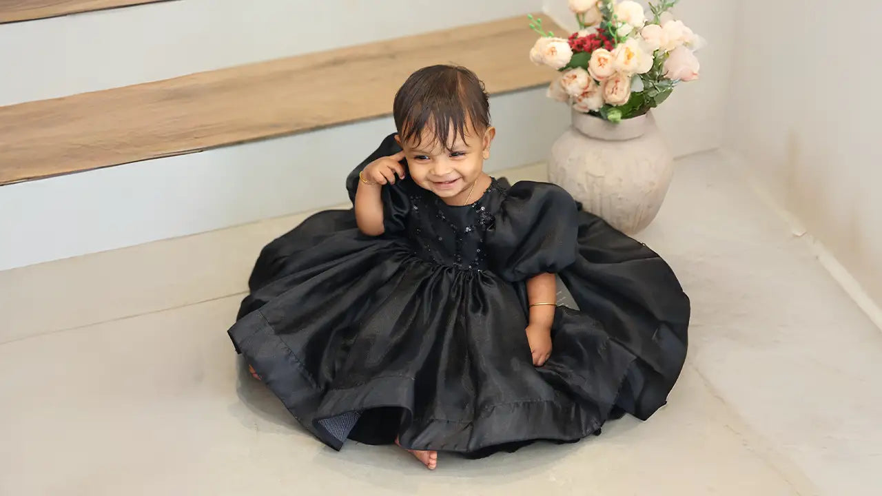Toddler in a black puff-sleeve party dress smiling while sitting indoors beside a vase of flowers