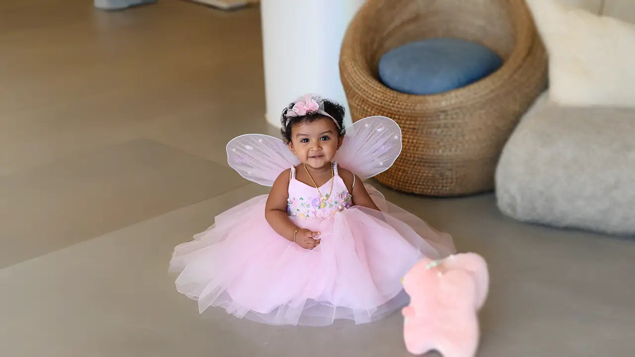 A baby girl wearing a voluminous pink soft net birthday frock with fairy wings and a matching headband.