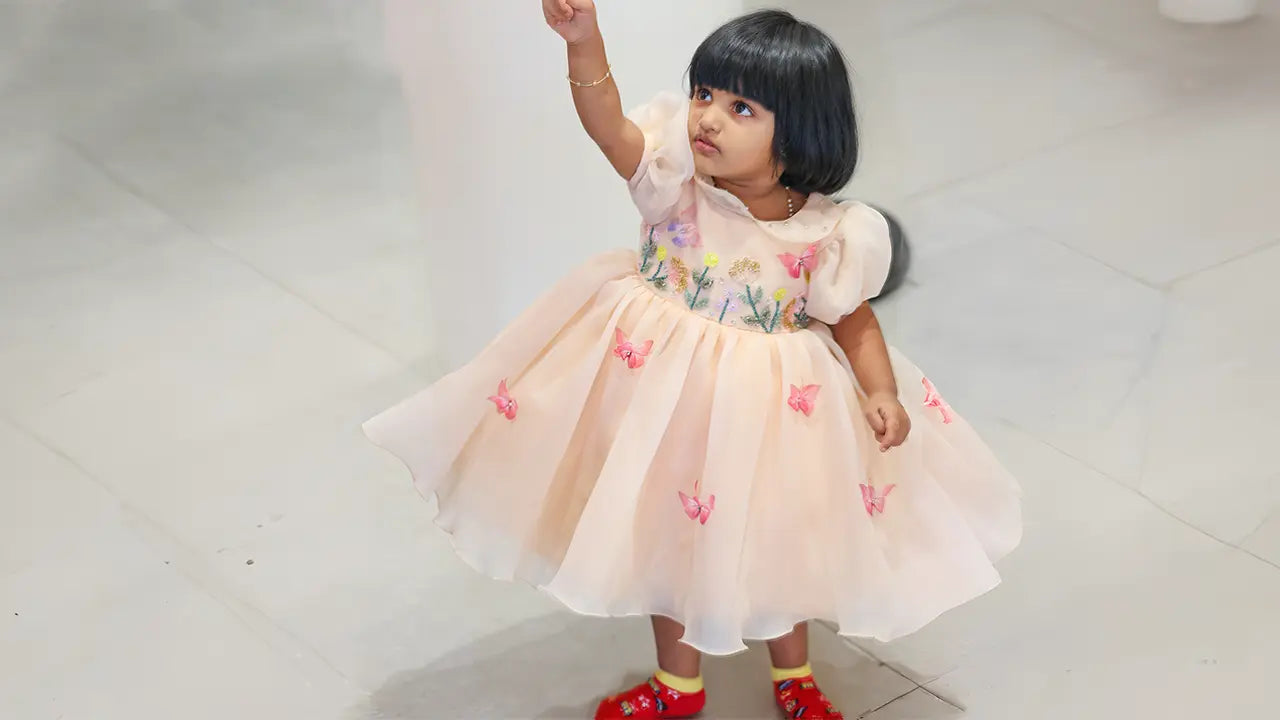 A little girl twirling in a peach-colored organza party wear frock adorned with realistic 3D butterfly appliqués and delicate hand-embroidered flower stems.