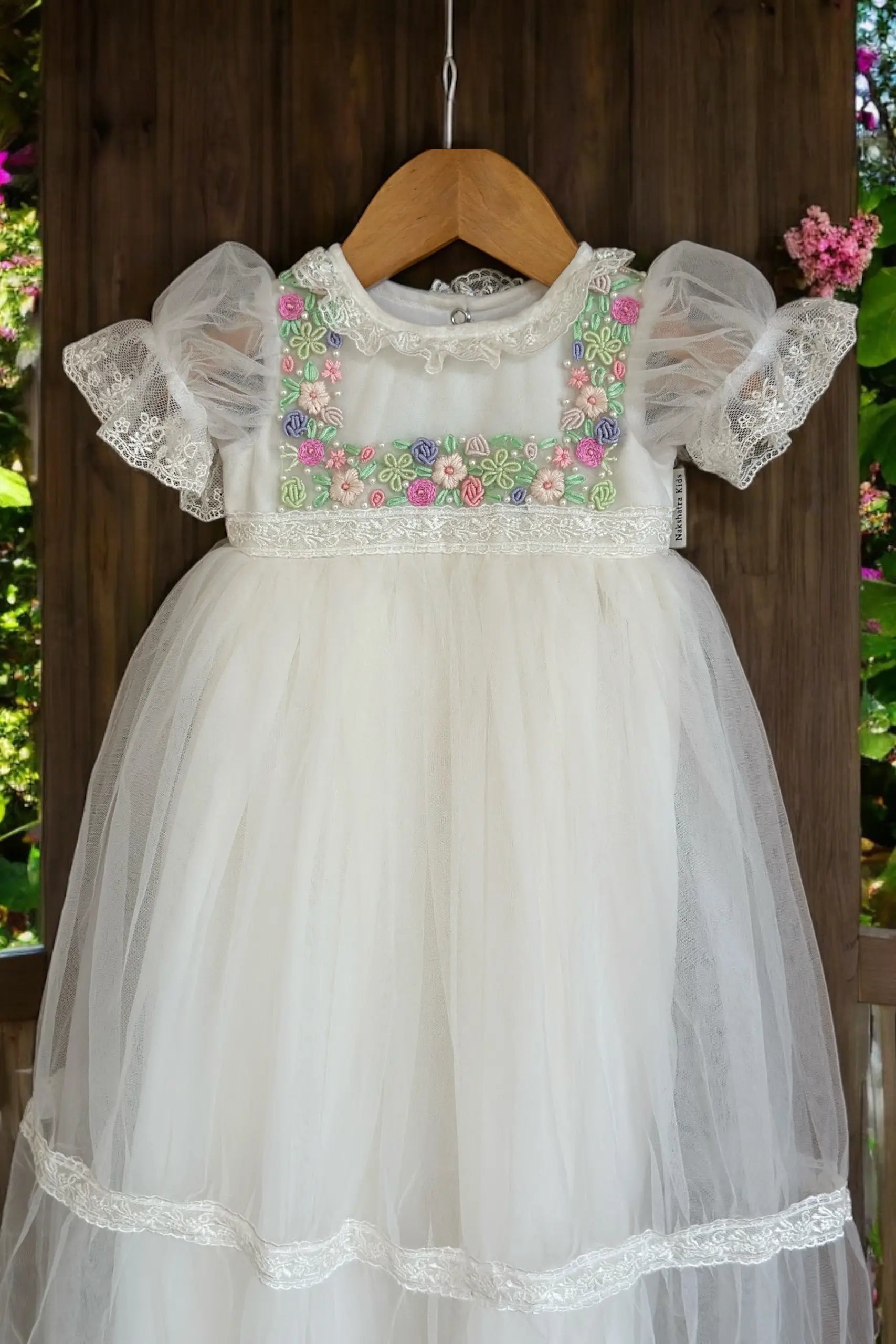Close view of an off-white lace dress with floral embroidery on a wooden hanger against a natural background.