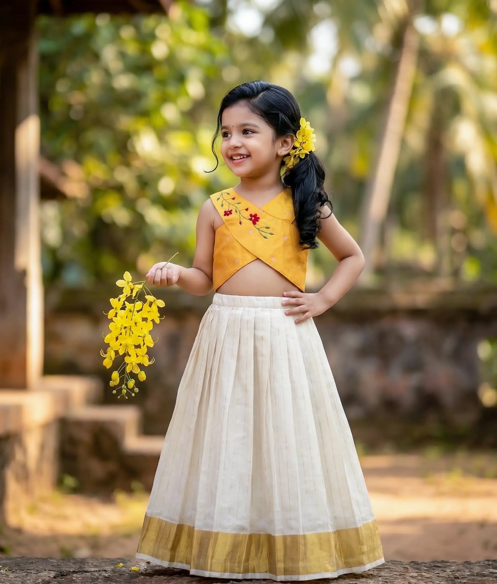 Baby girl wearing a yellow top with floral embroidery and a kasavu pleated skirt with vertical lines.