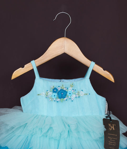 Close view of an sky blue tulle net frock with blue flowers and small pearls, displayed on a wooden hanger.