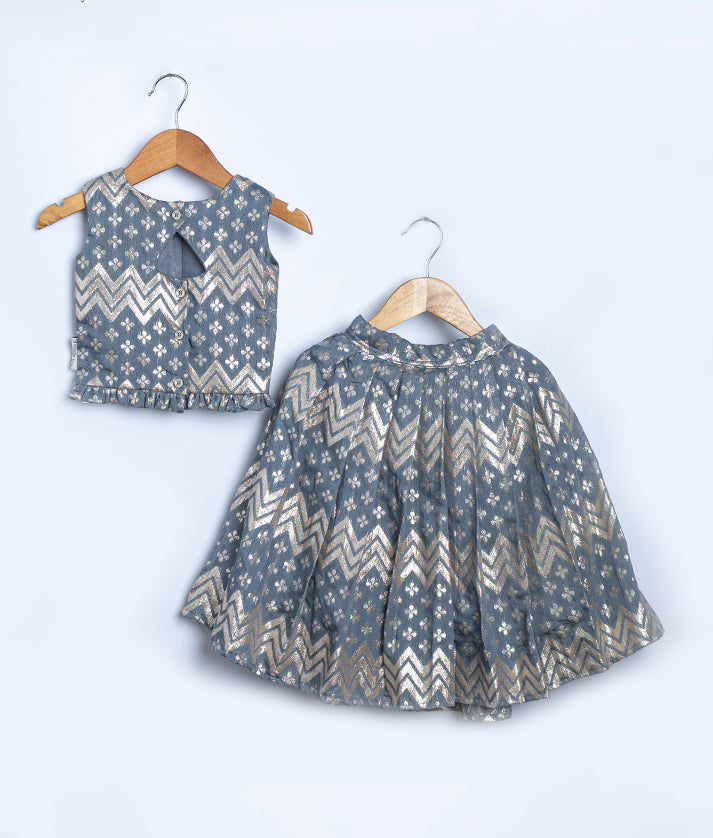 Back view of a stylish kid's ethnic outfit displayed against a light background, featuring a sleeveless blue-grey blouse with gold zigzag and floral patterns, back button closure, and a keyhole detail. Paired with a matching flared skirt with the same shimmering design, both pieces are hung on wooden hangers.
