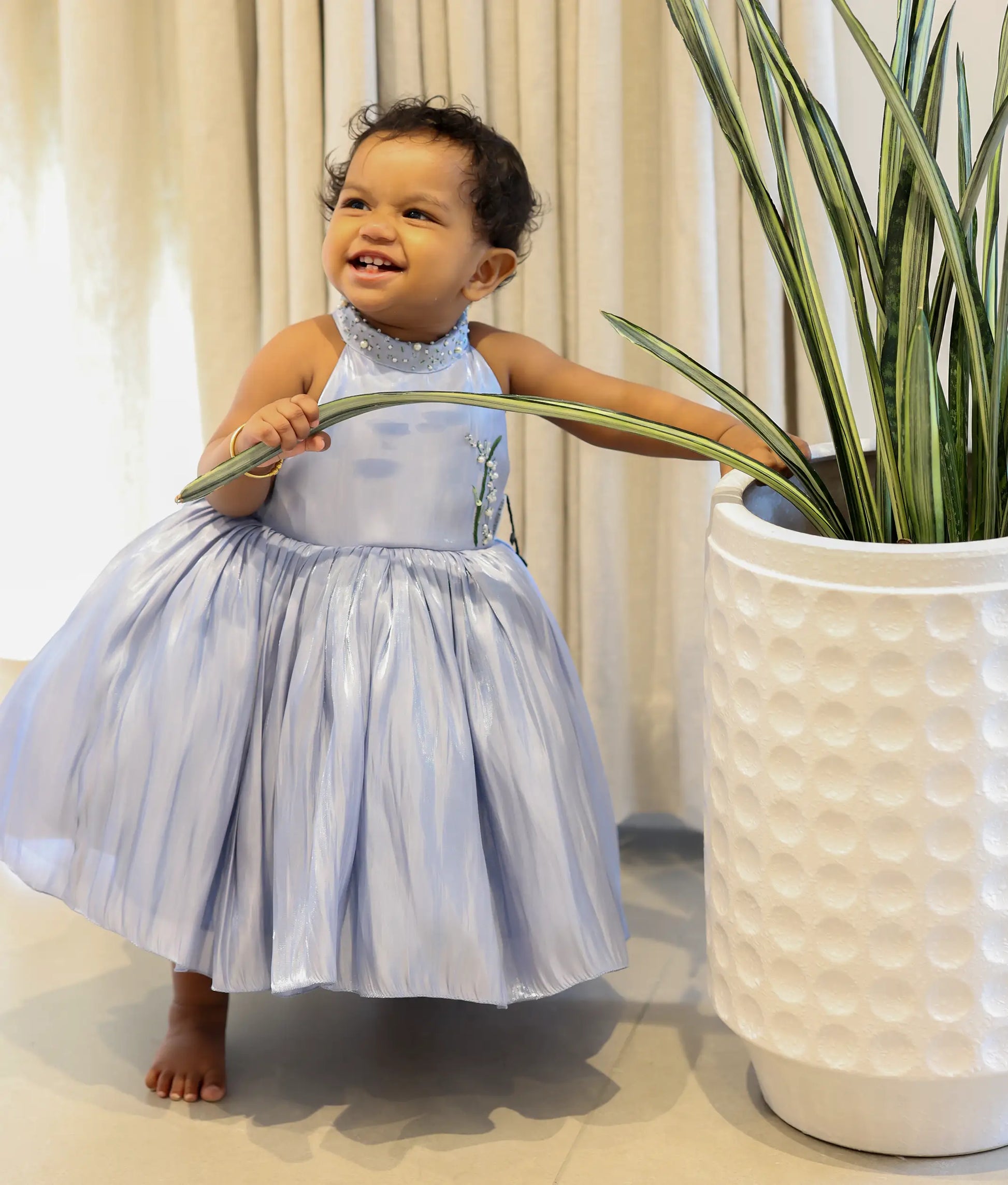 Baby girl wearing an ash blue laser organza floral handworked birthday frock for baby girls featuring a halter-neck and pintuck pleats