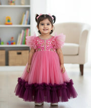 A baby girl wearing a pink handworked tulle net birthday gown with butterfly sleeves and a full-length flared bottom.