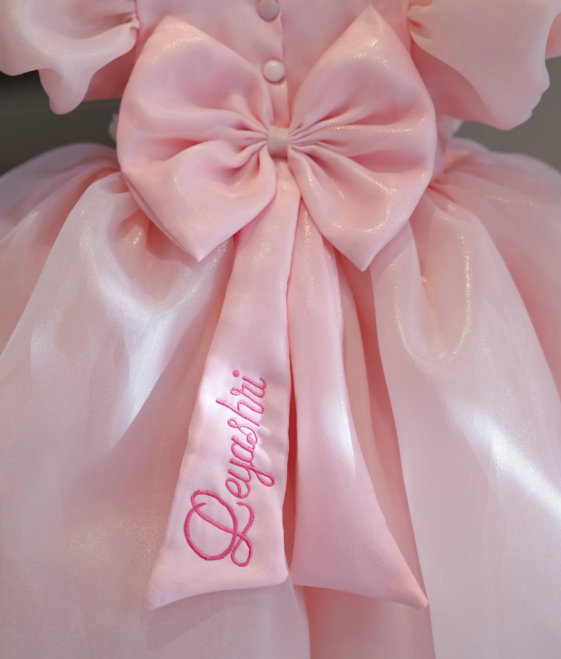 Close-up of the giant back bow on the baby pink shimmer organza dress, showcasing the customized pink name embroidery "Deyashri" on the ribbon tail.
