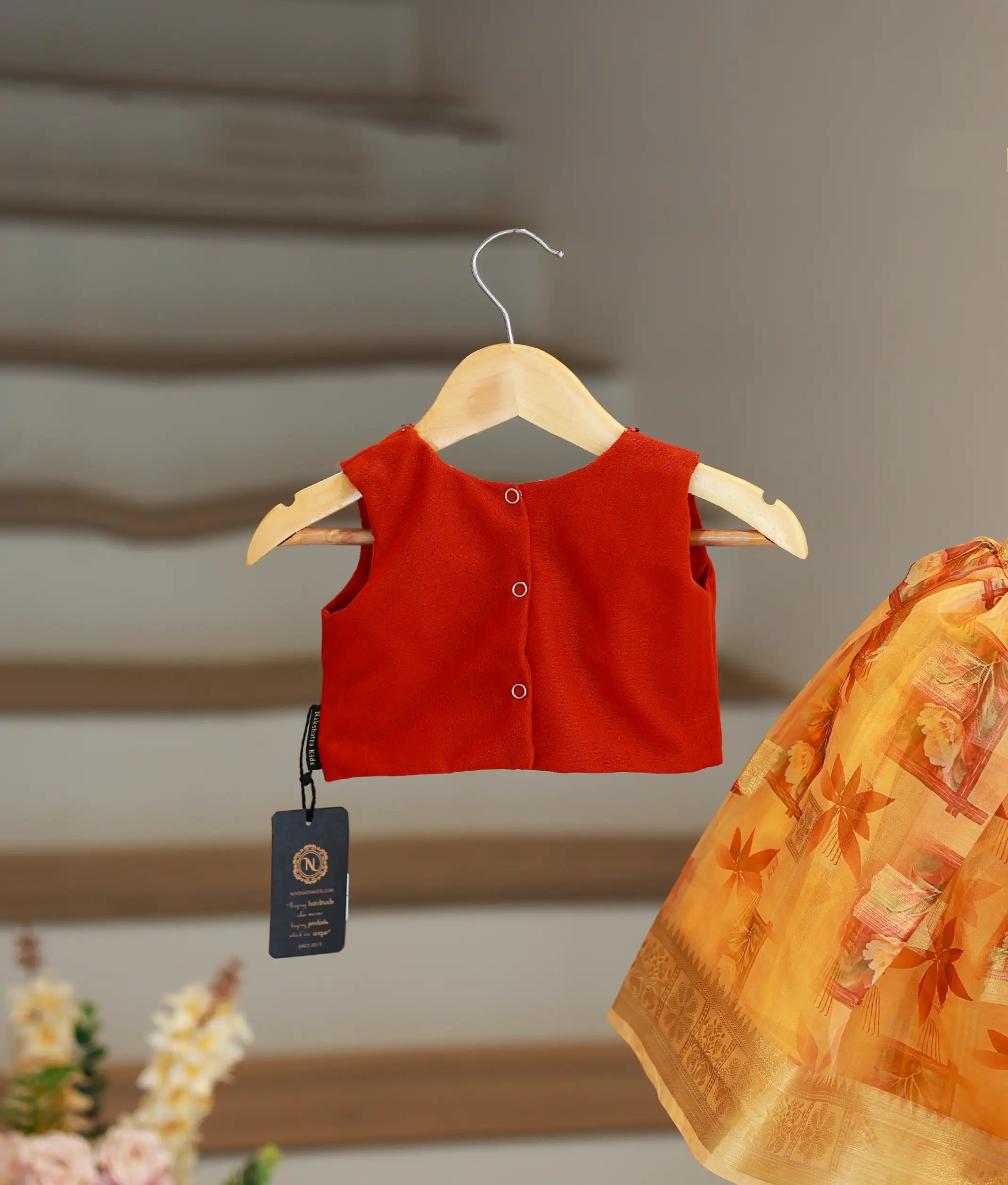 The back view of the red top showing the square V-neckline and press-button closure, hanging next to the yellow floral skirt