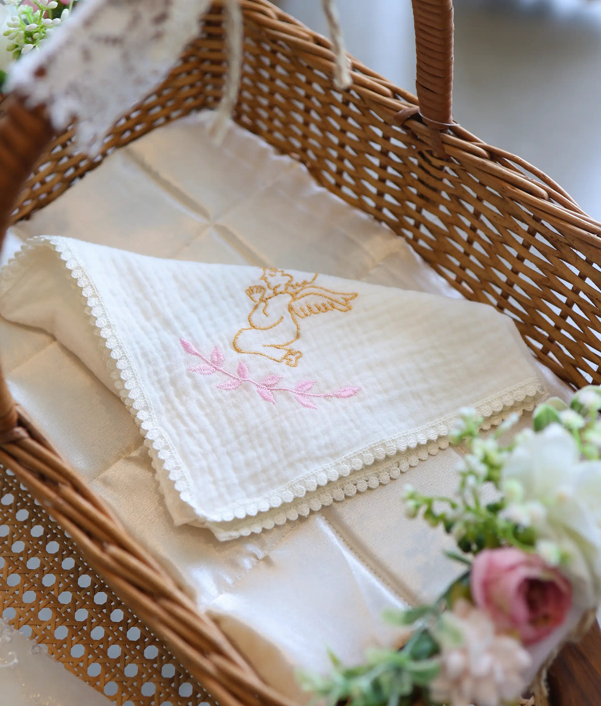Soft off-white baptism towel with embroidered angel motif neatly folded inside a floral-adorned woven basket.