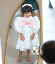 A little girl modeling a premium off-white embroidered party frock in a mirror reflection, showcasing the large pastel pink organza chest bow.