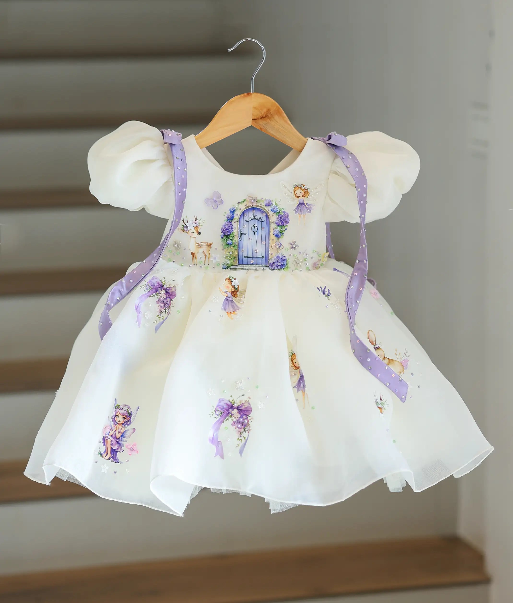 Full front view of a premium kid's off-white organza fairy-theme party frock on a hanger, showcasing the printed fairy garden design and flowing lavender shoulder ribbons.