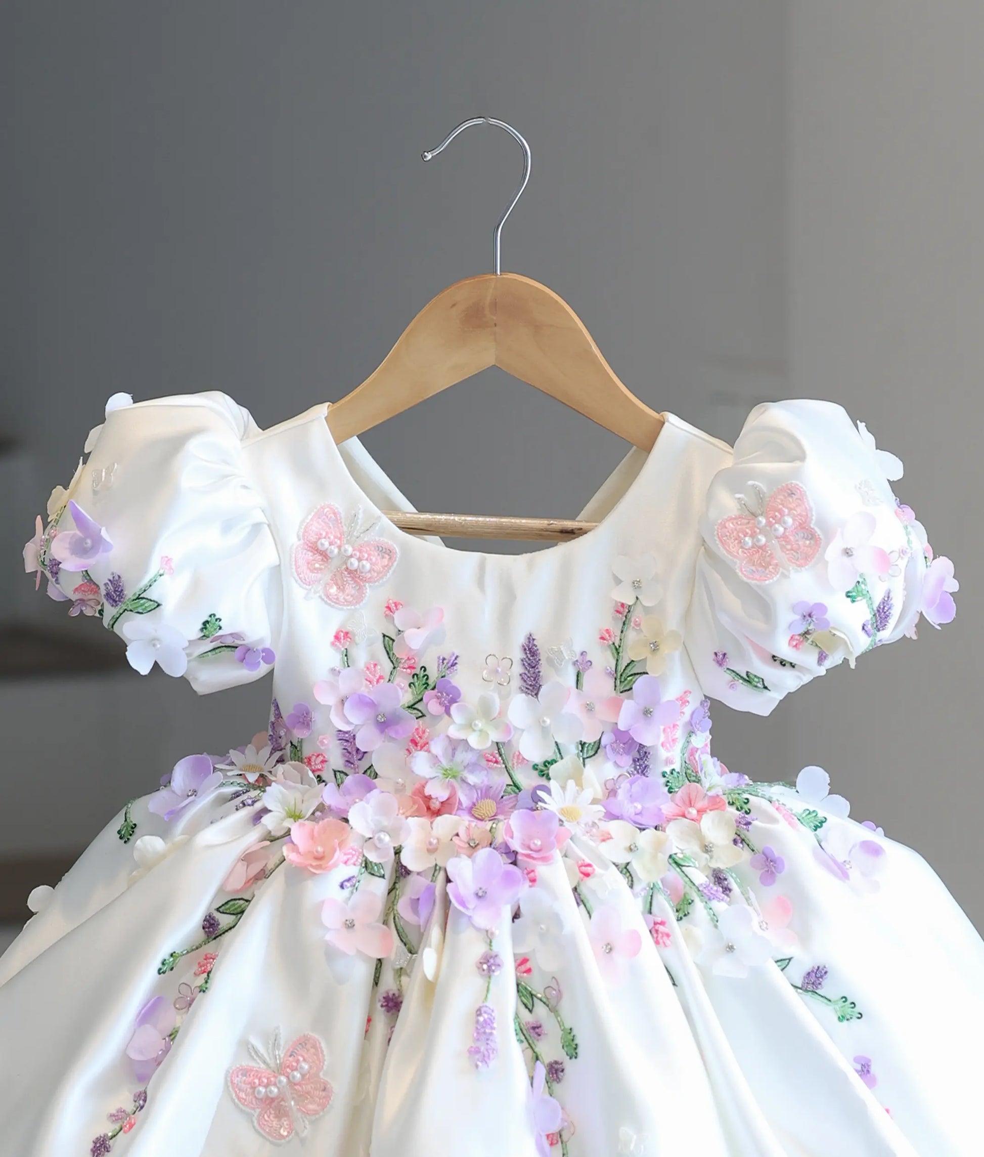 Close-up front view of the kid's dress bodice, showing the detailed 3D fabric flowers in lilac and peach, puff sleeves, and embroidered pink lace butterflies.