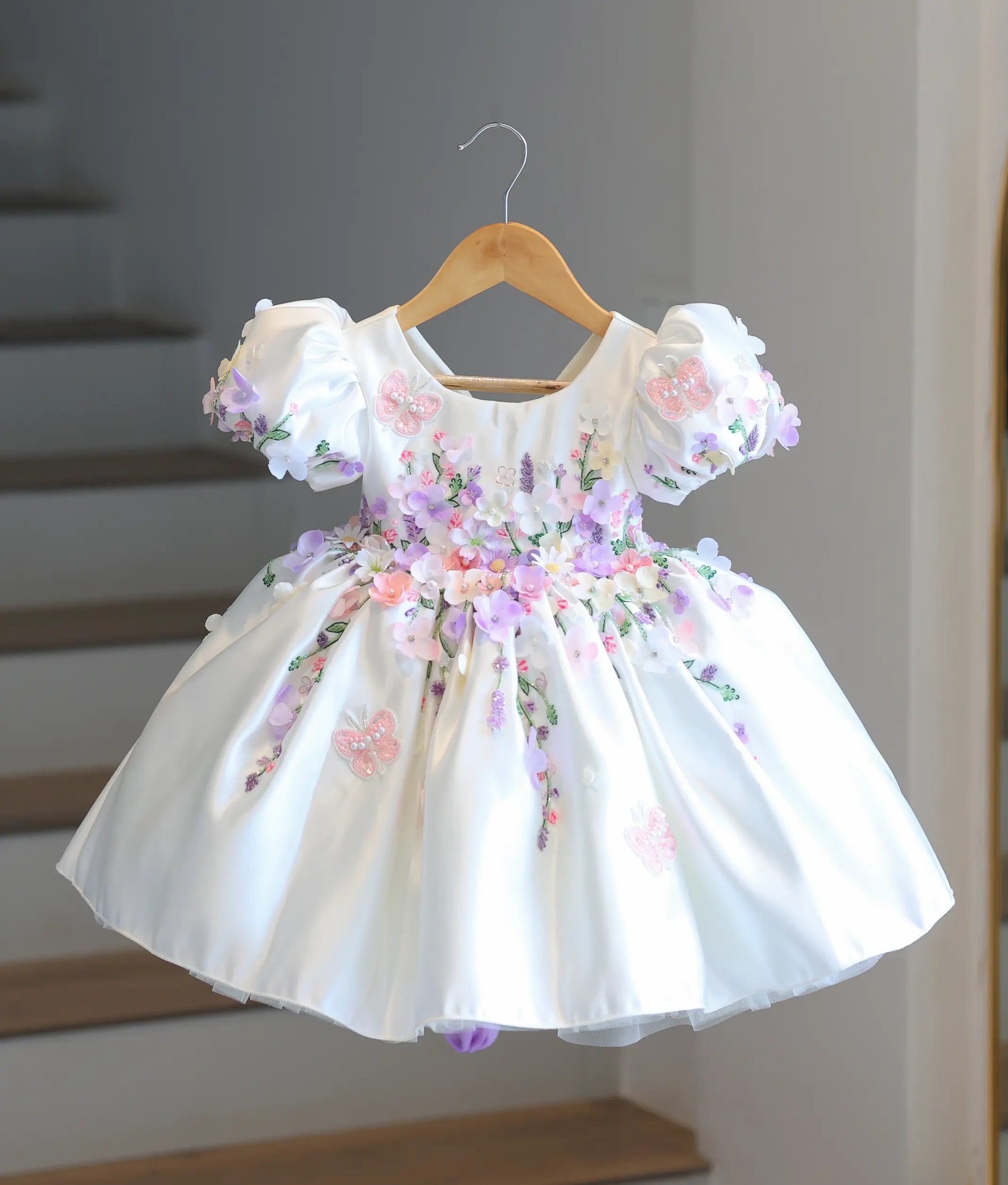 Full front view of a premium off-white satin kids' party frock hanging on a wooden hanger, covered in dense 3D pastel flowers and embroidered butterflies.