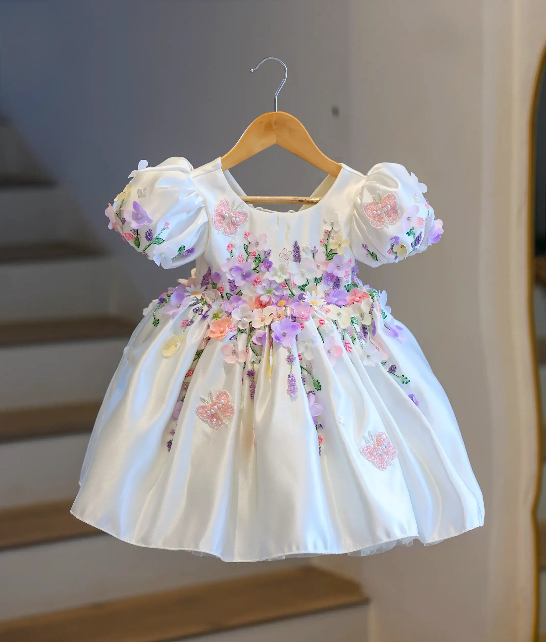 Full front view of a premium off-white satin kids' party frock hanging on a wooden hanger, covered in dense 3D pastel flowers and embroidered butterflies.