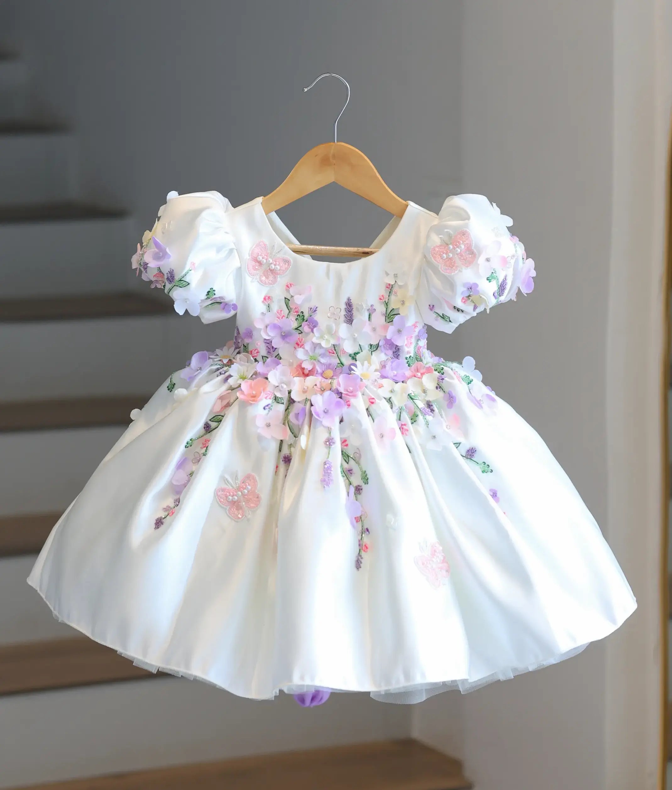 Full front view of a premium off-white satin kids' party frock hanging on a wooden hanger, covered in dense 3D pastel flowers and embroidered butterflies.