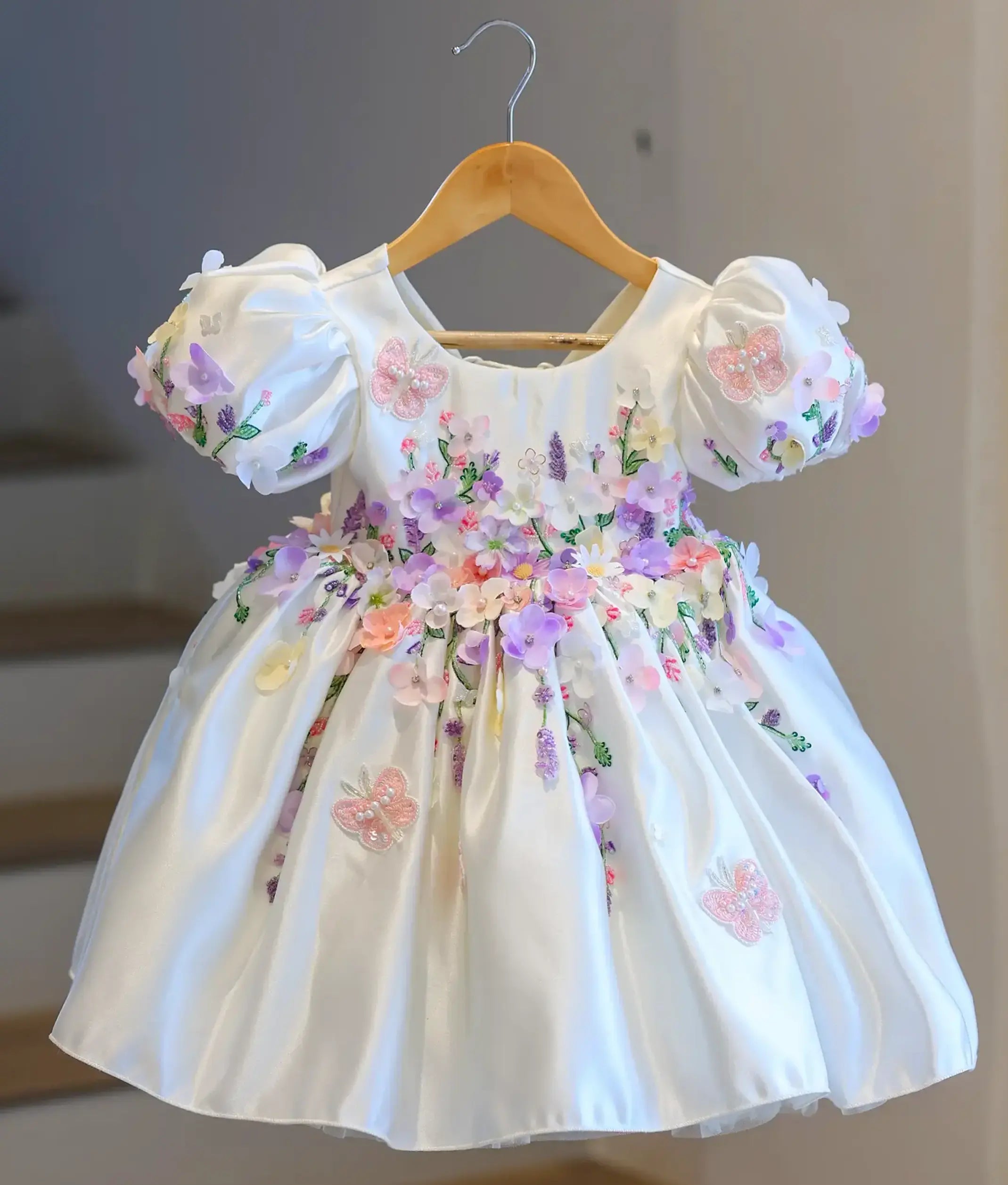 Full front view of a premium off-white satin kids' party frock hanging on a wooden hanger, covered in dense 3D pastel flowers and embroidered butterflies.