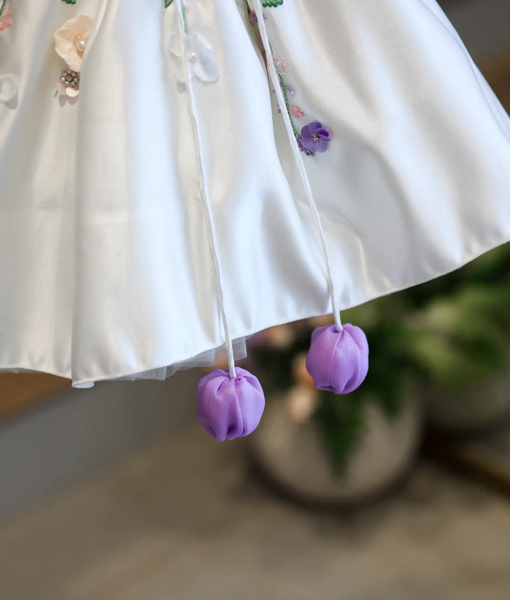 Detailed close-up of the dress hemline, showcasing the smooth off-white satin edge and two dangling bright purple fabric tulip tassels.