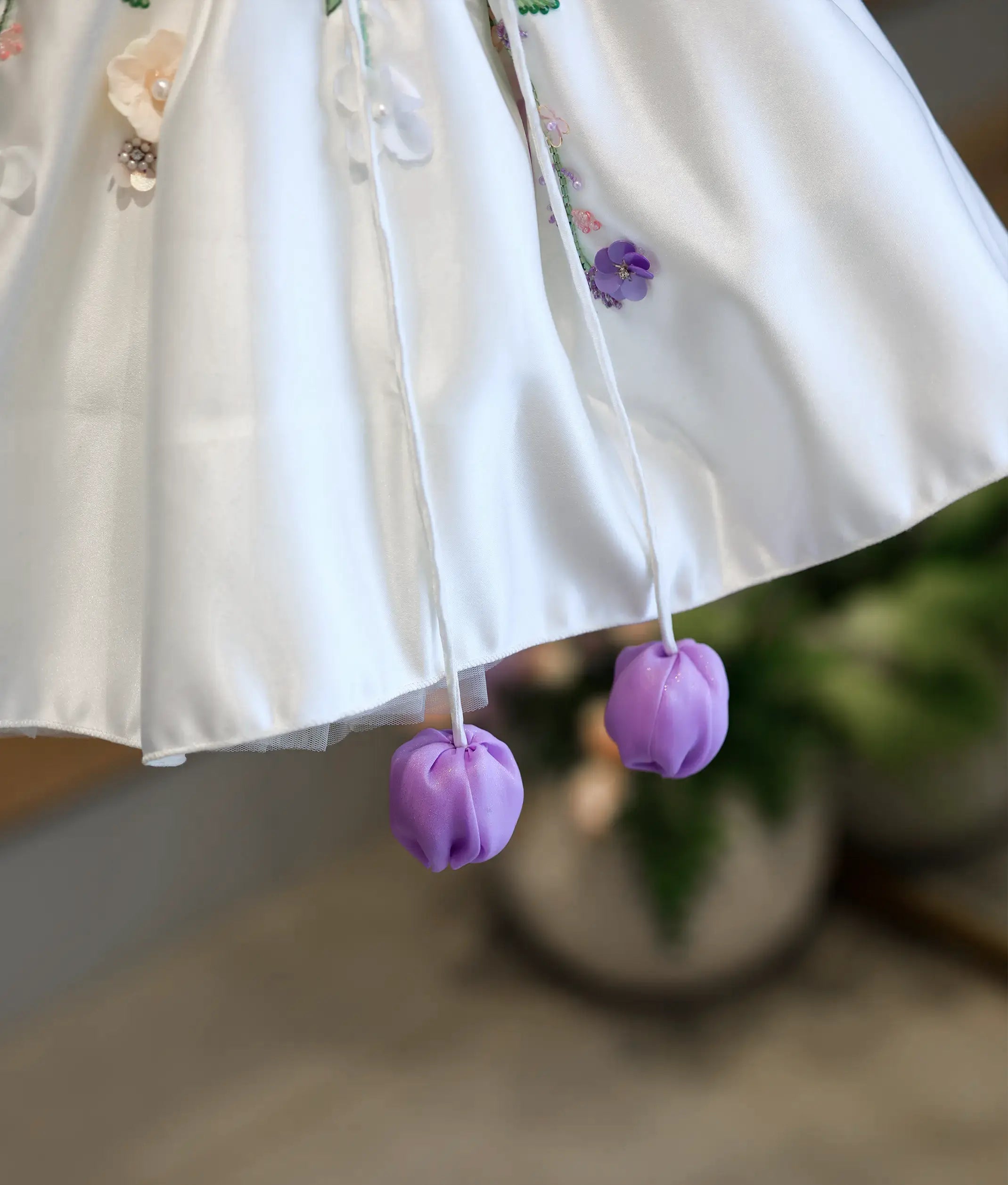 Detailed close-up of the dress hemline, showcasing the smooth off-white satin edge and two dangling bright purple fabric tulip tassels.
