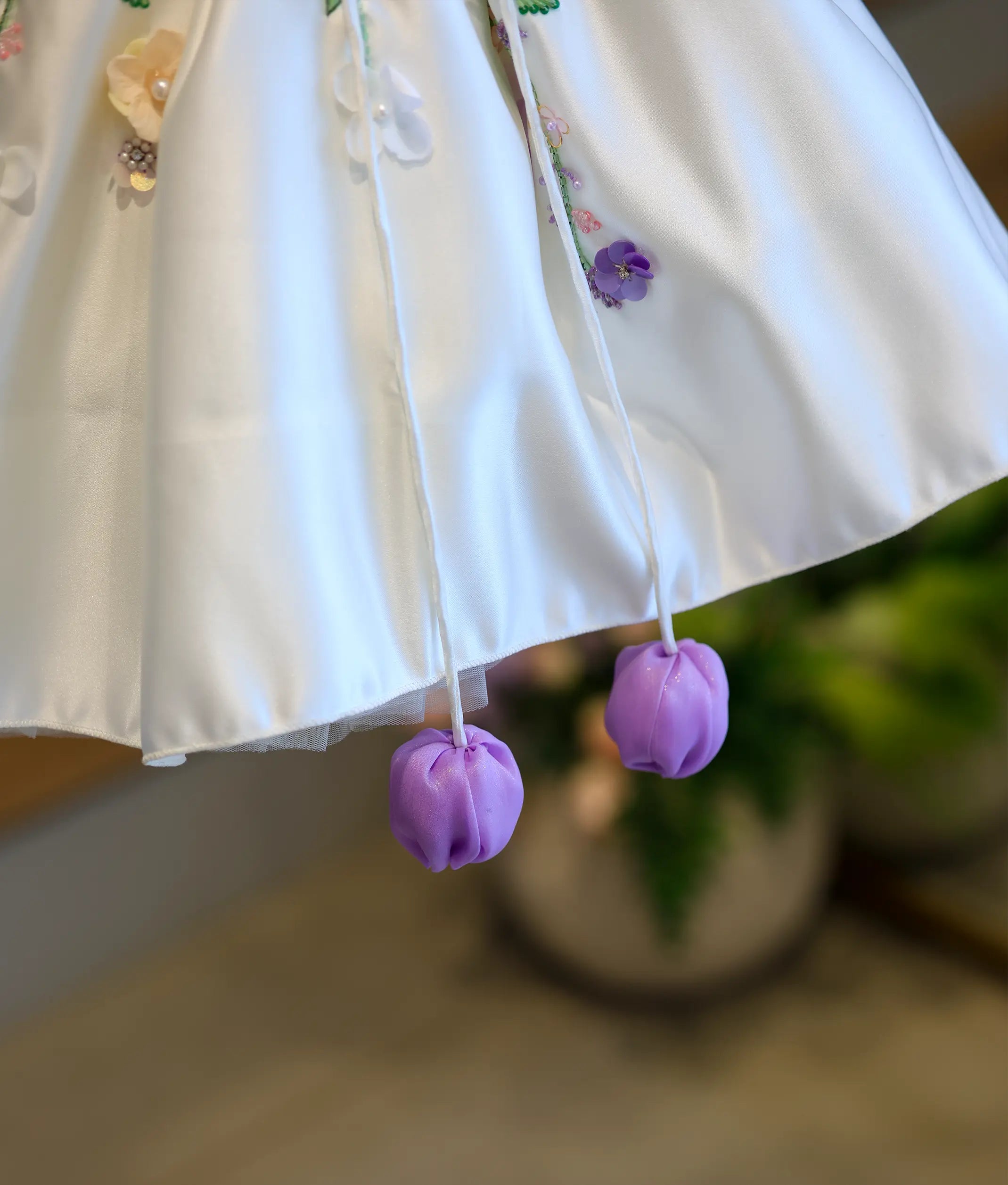 Detailed close-up of the dress hemline, showcasing the smooth off-white satin edge and two dangling bright lavender fabric tulip tassels.
