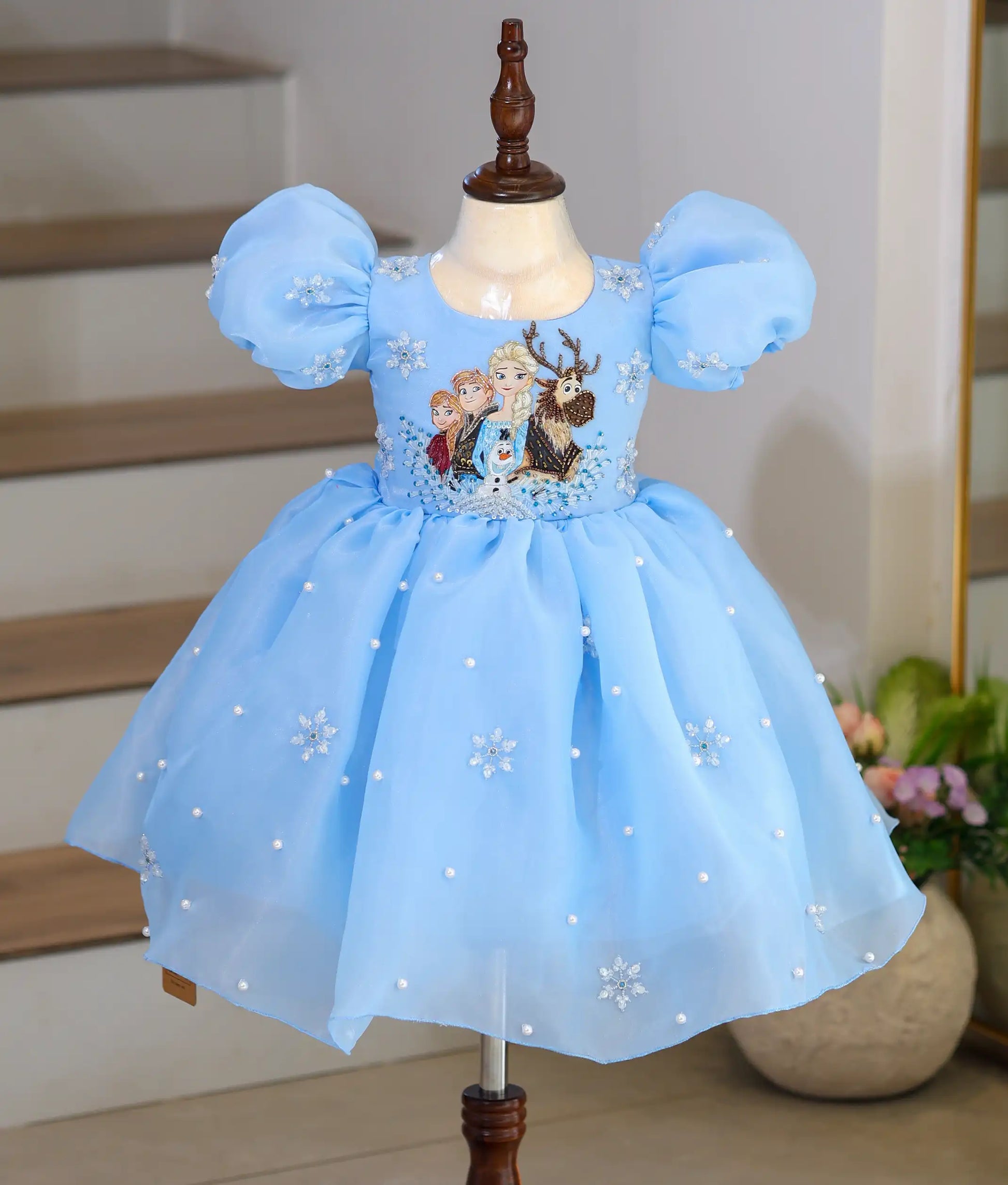 Frozen theme sky blue organza birthday frock for baby girl with hand embroidered characters, puff sleeves, pearl embellishments, and snowflake details front view.